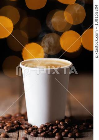 Steaming hot coffee in plain white paper cup on wooden table, with scattered roasted coffee beans and warm bokeh lights in the dark background creating cozy and inviting mood Steaming hot coffee in plain white paper cup on wooden table, with scattered roasted coffee beans and warm bokeh lights in the dark background creating cozy and inviting mood 139158804
