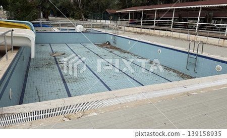 Abandoned outdoor swimming pool with debris and dry leaves, surrounded by a tiled deck Abandoned outdoor swimming pool with debris and dry leaves, surrounded by a tiled deck 139158935