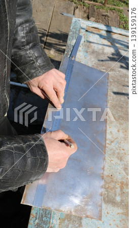 Marking out and measuring the required dimensions on a small sheet of metal lying on a work surface in an outdoor craft workshop, using a pencil and ruler in the hands of a craftsman marking out Marking out and measuring the required dimensions on a small sheet of metal lying on a work surface in an outdoor craft workshop, using a pencil and ruler in the hands of a craftsman marking out 139159176
