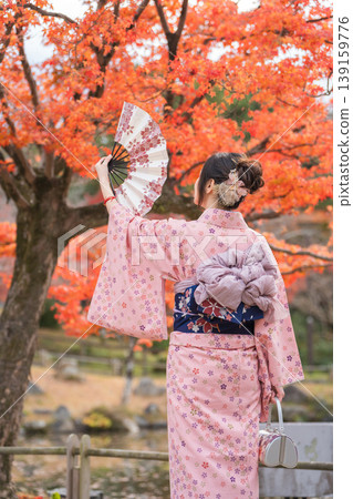 一位身穿浴衣（夏季和服）的女子手持扇子，背對著鏡頭，站在秋葉環繞的背景下。日本京都府。 139159776