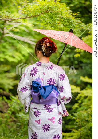 A woman in a yukata (summer kimono) stands with a red Japanese umbrella in a garden surrounded by fresh greenery, seen from behind. Kyoto Prefecture, Japan. A woman in a yukata (summer kimono) stands with a red Japanese umbrella in a garden surrounded by fresh greenery, seen from behind. Kyoto Prefecture, Japan. 139159777