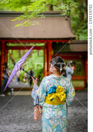 A woman in a yukata (summer kimono) walks from behind in the grounds of a shrine surrounded by fresh greenery, holding a purple Japanese umbrella. Kyoto Prefecture, Japan. A woman in a yukata (summer kimono) walks from behind in the grounds of a shrine surrounded by fresh greenery, holding a purple Japanese umbrella. Kyoto Prefecture, Japan. 139159782