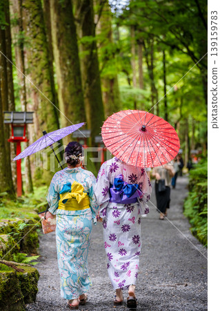 Two women in yukata (summer kimonos) walk along a vermilion-colored path lined with lanterns, carrying traditional Japanese umbrellas. Kyoto Prefecture, Japan Two women in yukata (summer kimonos) walk along a vermilion-colored path lined with lanterns, carrying traditional Japanese umbrellas. Kyoto Prefecture, Japan 139159783