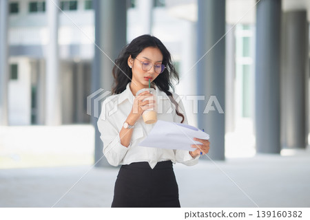 Young office professional drinking iced coffee and reading documents outdoors, showing focused work, daily routine, and modern business lifestyle in a bright corporate architectural setting Young office professional drinking iced coffee and reading documents outdoors, showing focused work, daily routine, and modern business lifestyle in a bright corporate architectural setting 139160382