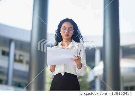 Low angle view of a businesswoman reviewing paperwork outside a modern building, conveying professionalism, independence, and active corporate workflow in natural daylight setting. Low angle view of a businesswoman reviewing paperwork outside a modern building, conveying professionalism, independence, and active corporate workflow in natural daylight setting. 139160434