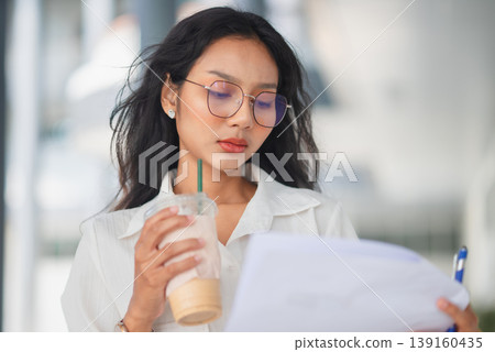 Close-up of a serious businesswoman in glasses reviewing paperwork with iced coffee in hand, emphasizing focus, analysis, and thoughtful decision making in a modern workplace environment. Close-up of a serious businesswoman in glasses reviewing paperwork with iced coffee in hand, emphasizing focus, analysis, and thoughtful decision making in a modern workplace environment. 139160435