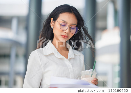 Stylish businesswoman in glasses reviewing papers and holding iced coffee in front of a modern office, illustrating thoughtful analysis, workplace focus, and professional urban lifestyle. 139160460