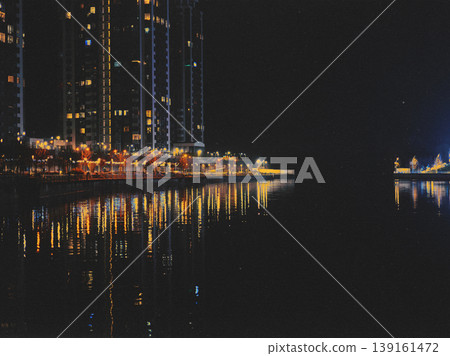 A scenic night view of a modern urban waterfront, illuminated high-rise residential buildings reflected in dark water. Nighttime cityscape. A scenic night view of a modern urban waterfront, illuminated high-rise residential buildings reflected in dark water. Nighttime cityscape. 139161472