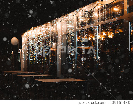 Cozy winter evening ambiance at an outdoor cafe terrace during snowfall. The scene is illuminated by glowing string fairy lights. Cafe patio, terrace. Cozy winter evening ambiance at an outdoor cafe terrace during snowfall. The scene is illuminated by glowing string fairy lights. Cafe patio, terrace. 139161488