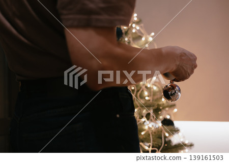 A person's hands decorating a small Christmas tree indoors with ornaments and lights, holiday preparation at home. Family tradition and culture. 139161503