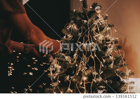 Close-up of female hands decorating a Christmas tree with a shiny silver bauble and string lights. Authentic holiday cozy scene. Led garland lights Close-up of female hands decorating a Christmas tree with a shiny silver bauble and string lights. Authentic holiday cozy scene. Led garland lights 139161512