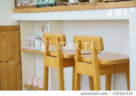 Cozy kitchen corner with wooden bar stools, white tiled wall, and decorative ceramic jars on open shelves in a warm home environment Cozy kitchen corner with wooden bar stools, white tiled wall, and decorative ceramic jars on open shelves in a warm home environment 139162316