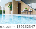 Modern indoor swimming pool with wooden accents, white chairs, large potted plant, glass windows, and blue-tiled pool for relaxing leisure 139162347