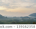 Scenic landscape of rolling green hills with misty fog and distant mountains under a cloudy sky during early morning sunrise 139162358