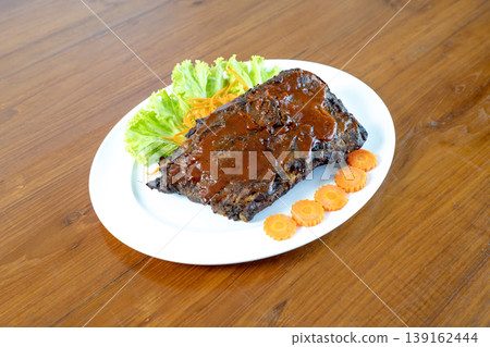 Roasted BBQ pork ribs glazed with savory sauce served on white plate with lettuce and carrot garnish on wooden table. Delicious grilled meat dish for restaurant menu or food blogging. 139162444