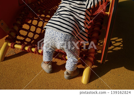 A one-year-old child playing while holding onto something to stand. A toddler's back view playing on playground equipment. A one-year-old child playing while holding onto something to stand. A toddler's back view playing on playground equipment. 139162824