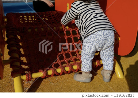 A one-year-old child playing while holding onto something to stand. A toddler's back view playing on playground equipment. A one-year-old child playing while holding onto something to stand. A toddler's back view playing on playground equipment. 139162825