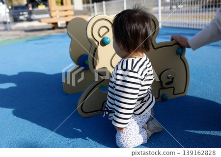 一個一歲大的孩子在公園的遊樂設施上玩耍，這是他的背影。人行道是藍色的。 139162828