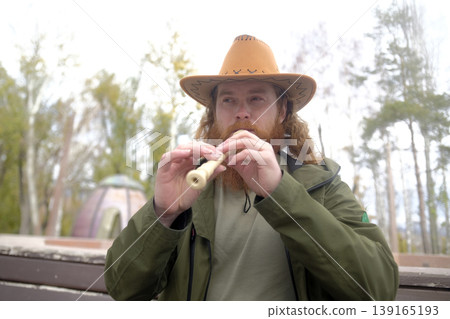 A red-haired man with curly hair and a beard is playing a flute. A musician in a cowboy hat plays a pipe in the park. The image is made in a rustic and peaceful style 139165193