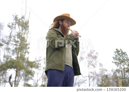 A handsome red-bearded man in a cowboy hat is playing the flute in the park. The piper plays the pipe, bottom view. Portrait of a free musician dressed in a green jacket A handsome red-bearded man in a cowboy hat is playing the flute in the park. The piper plays the pipe, bottom view. Portrait of a free musician dressed in a green jacket 139165200