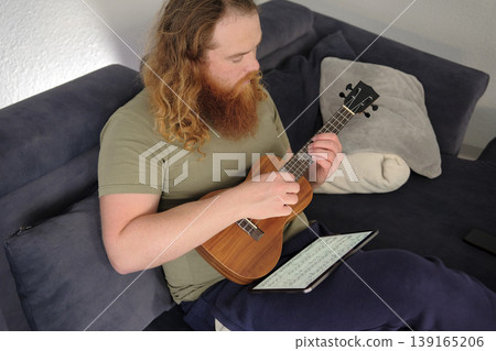 An authentic red-haired bearded man learns to play the ukulele from notes on a tablet. A man learns a new tune while sitting on the couch at home. A musician An authentic red-haired bearded man learns to play the ukulele from notes on a tablet. A man learns a new tune while sitting on the couch at home. A musician 139165206