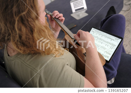 A man with long wavy red hair and a beard is learning to play the ukulele. A musician learns a new melody from notes on a tablet. The guitarist is sitting on the couch A man with long wavy red hair and a beard is learning to play the ukulele. A musician learns a new melody from notes on a tablet. The guitarist is sitting on the couch 139165207