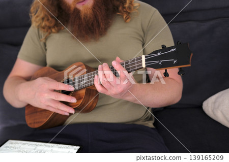 A red-haired guitarist learns to play the ukulele from notes from a tablet. A man with a beard and long hair is playing the ukulele sitting on the couch. The musician A red-haired guitarist learns to play the ukulele from notes from a tablet. A man with a beard and long hair is playing the ukulele sitting on the couch. The musician 139165209