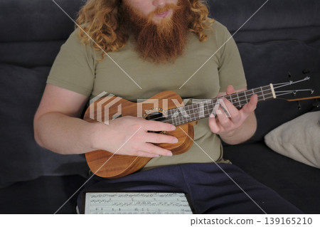Ukulele lessons. A man with curly red hair and a beard is playing the ukulele sitting on the couch. The guitarist learns a new melody from the notes on the tablet Ukulele lessons. A man with curly red hair and a beard is playing the ukulele sitting on the couch. The guitarist learns a new melody from the notes on the tablet 139165210