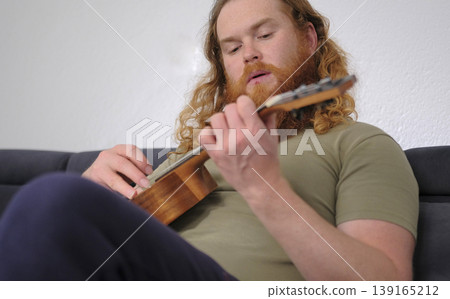 A man with a red curly beard and wavy red hair is playing the ukulele in his room. He is sitting on a blue sofa and carefully playing chords. The guitarist is learning a new tune A man with a red curly beard and wavy red hair is playing the ukulele in his room. He is sitting on a blue sofa and carefully playing chords. The guitarist is learning a new tune 139165212