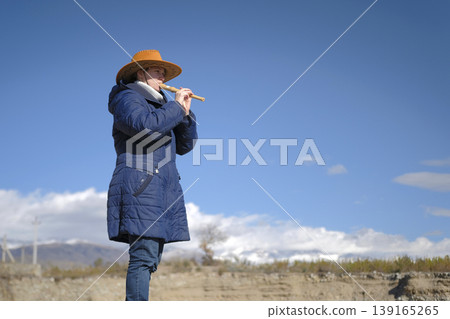 A cowboy woman in a wide-brimmed hat and a blue coat is playing a flute in a field. The sky is blue, the clouds are white, and mountains are visible in the background. A piper's concert in nature A cowboy woman in a wide-brimmed hat and a blue coat is playing a flute in a field. The sky is blue, the clouds are white, and mountains are visible in the background. A piper's concert in nature 139165265