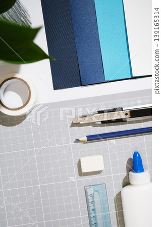 Craft supplies flat lay on cutting mat: utility knife, pencil, eraser, glue, tape, ruler, and blue cardstock sheets. Top view, white background. 139165314