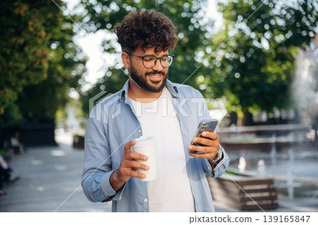 Cup of drink and smartphone are in hands. Handsome man is outdoors in the city Cup of drink and smartphone are in hands. Handsome man is outdoors in the city 139165847