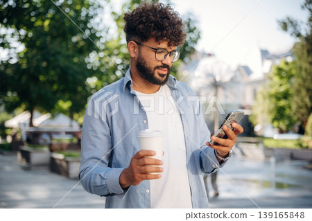 Paper cup in right hand and smartphone in left one. Handsome man is outdoors in the city Paper cup in right hand and smartphone in left one. Handsome man is outdoors in the city 139165848