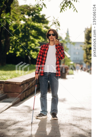 Smartphone in hand, talking. Blind young guy is in the town outdoors Smartphone in hand, talking. Blind young guy is in the town outdoors 139166154