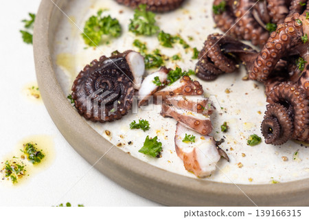 Boiled octopus tentacles and sliced pieces on ceramic plate closeup. Olive oil and parsley dressing 139166315