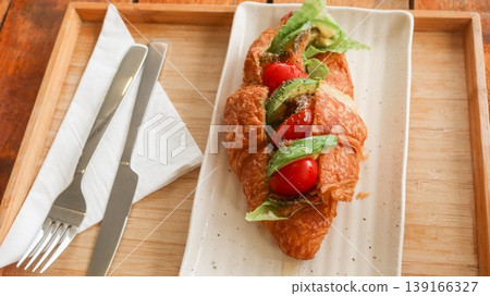 close up of a Golden croissant sandwich filled with avocado, cherry tomatoes, lettuce, and seasoning, served on white plate with cutlery on a wooden tray close up of a Golden croissant sandwich filled with avocado, cherry tomatoes, lettuce, and seasoning, served on white plate with cutlery on a wooden tray 139166327