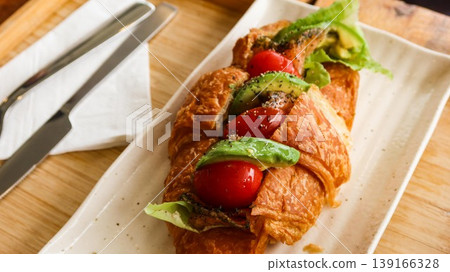 close up of a Golden croissant sandwich filled with avocado, cherry tomatoes, lettuce, and seasoning, served on white plate with cutlery on a wooden tray close up of a Golden croissant sandwich filled with avocado, cherry tomatoes, lettuce, and seasoning, served on white plate with cutlery on a wooden tray 139166328