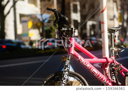 A pink bicycle is parked on a pole on a city street, secured with a chain lock. A pink bicycle is parked on a pole on a city street, secured with a chain lock. 139166361