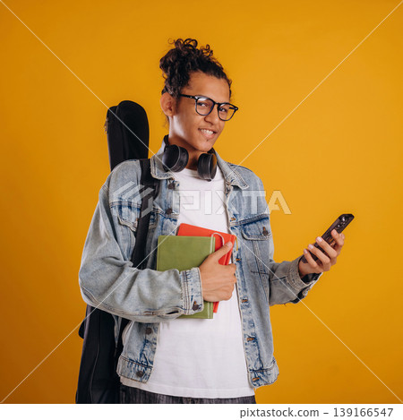 Guitar backpack on back and notepads in hand. Young guy is against yellow background Guitar backpack on back and notepads in hand. Young guy is against yellow background 139166547