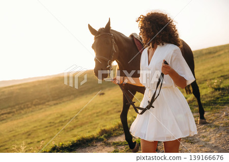 The walk on the footpath. Woman in white dress is with horse on the countryside field The walk on the footpath. Woman in white dress is with horse on the countryside field 139166676
