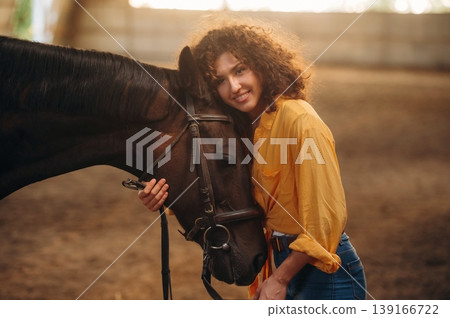 Yellow colored natural sunlight, standing and posing. Beautiful young woman is with horse in hangar Yellow colored natural sunlight, standing and posing. Beautiful young woman is with horse in hangar 139166722