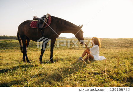 Sitting on the ground, side view. Woman in white dress is with horse on the countryside field Sitting on the ground, side view. Woman in white dress is with horse on the countryside field 139166723