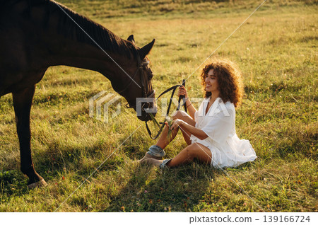 Sitting on the ground, side view. Woman in white dress is with horse on the countryside field Sitting on the ground, side view. Woman in white dress is with horse on the countryside field 139166724