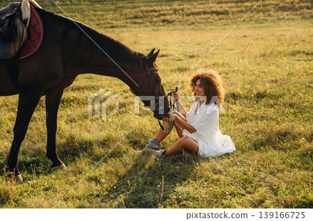 Sitting on the ground, side view. Woman in white dress is with horse on the countryside field Sitting on the ground, side view. Woman in white dress is with horse on the countryside field 139166725