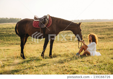 Sitting on the ground, side view. Woman in white dress is with horse on the countryside field Sitting on the ground, side view. Woman in white dress is with horse on the countryside field 139166726
