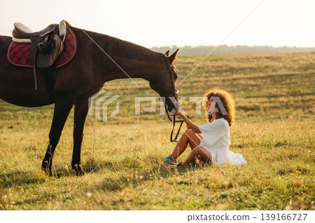 Sitting on the ground, side view. Woman in white dress is with horse on the countryside field Sitting on the ground, side view. Woman in white dress is with horse on the countryside field 139166727