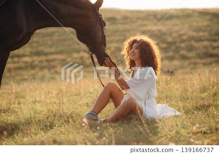 Side view, sitting on the ground. Woman in white dress is with horse on the countryside field Side view, sitting on the ground. Woman in white dress is with horse on the countryside field 139166737