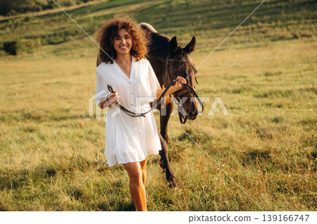 Front view, posing. Woman in white dress is with horse on the countryside field Front view, posing. Woman in white dress is with horse on the countryside field 139166747