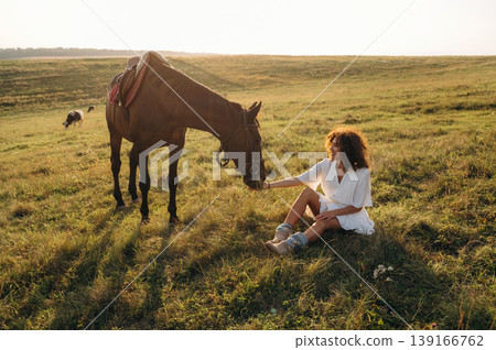 Resting, sitting on the ground. Woman in white dress is with horse on the countryside field Resting, sitting on the ground. Woman in white dress is with horse on the countryside field 139166762