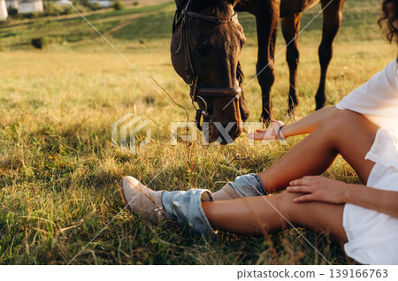 Resting, sitting on the ground. Woman in white dress is with horse on the countryside field Resting, sitting on the ground. Woman in white dress is with horse on the countryside field 139166763
